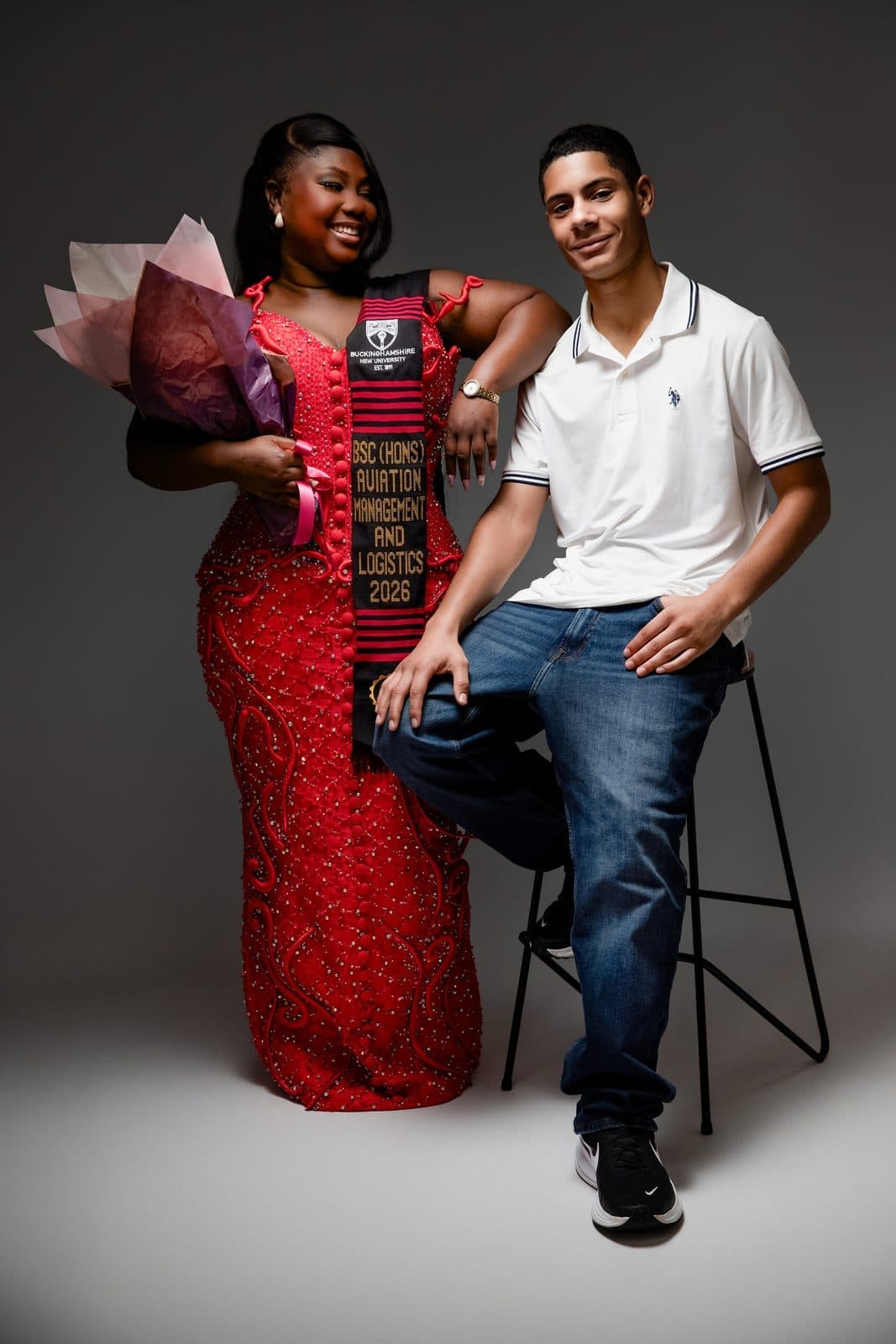 Graduate in red gown and kente stole with a young man seated on a stool
