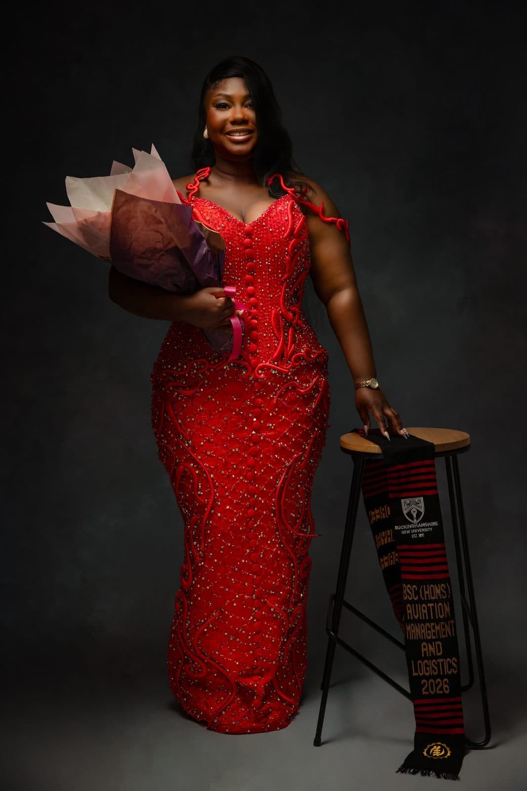 Graduate in red beaded gown with kente stole and bouquet, studio portrait